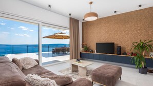 A modern living room with a large sofa, a coffee table, and a TV on a wooden console. There is a balcony with a view of the sea.