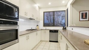 A modern kitchen with white cabinets, a built-in oven, a dishwasher, and a window with a view of trees.