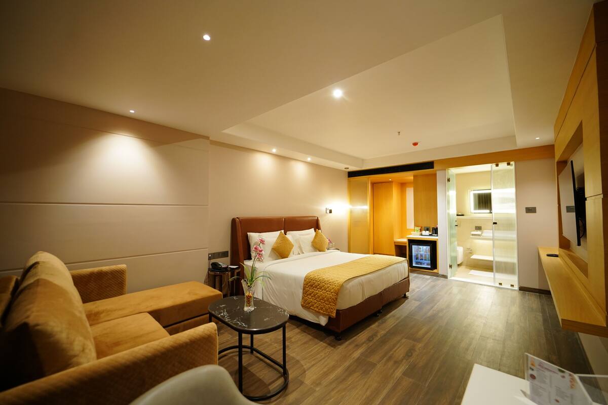 A hotel room with a bed, a sofa, a small table with a vase, and a TV.