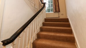 Een trap met witte balustrades en een zwarte leuning, die uitkomt op een plateau met een radiator en een raam met gordijnen met een patroon.