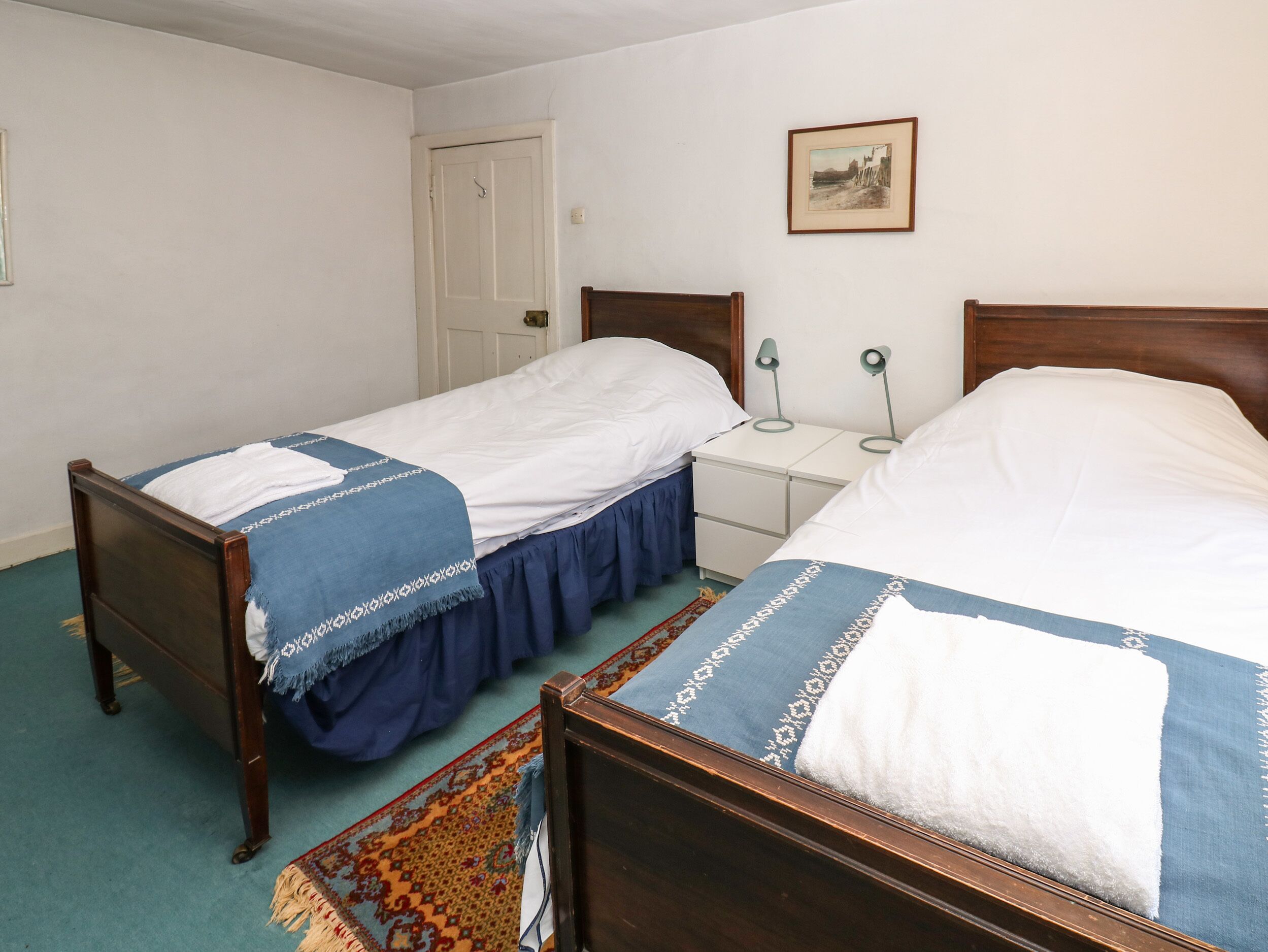 A room with two wooden beds, a white door, a small bedside table with a lamp, and a framed picture on the wall.