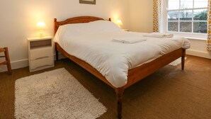 Een keurig opgemaakt bed in een kamer met een houten hoofdbord, een witte nachtkastje met een lamp, een kleedje en een raam met gordijnen met een patroon.