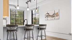 A modern kitchen with three bar stools, a wooden table, and a large window with a view of trees.