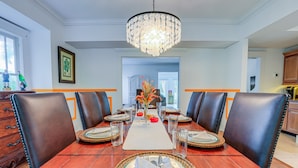 A dining room with a wooden table set for six, surrounded by leather chairs, a chandelier, and a vase with flowers.