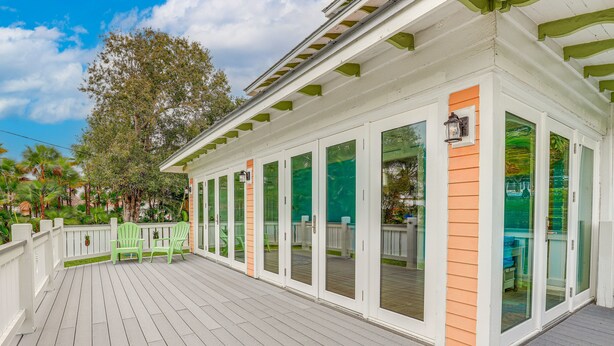 A covered deck with white railings and green chairs, overlooking a backyard with a wooden deck and a large tree.