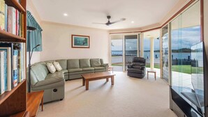 A living room with a sectional sofa, a coffee table, a bookshelf, a black leather chair, and large windows offering a view of the outdoors.