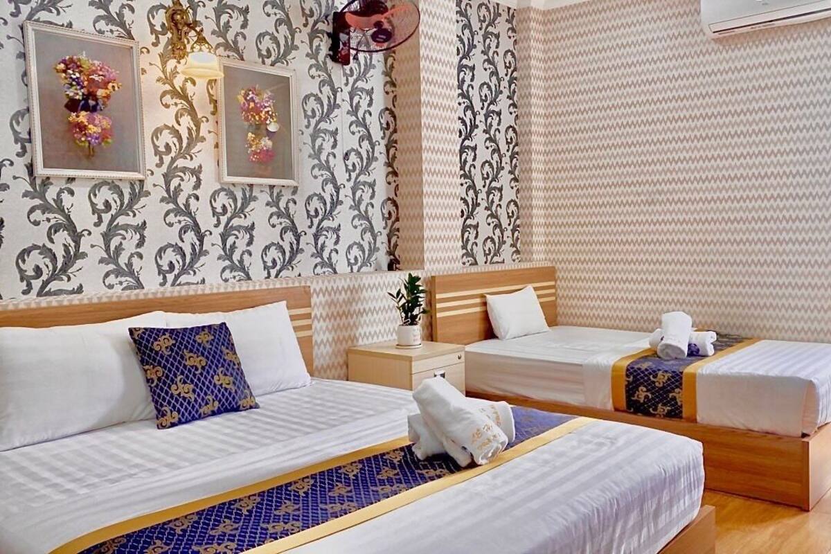 A hotel room with two beds, a small table, and a chair. There are decorative wall panels and a window with curtains.