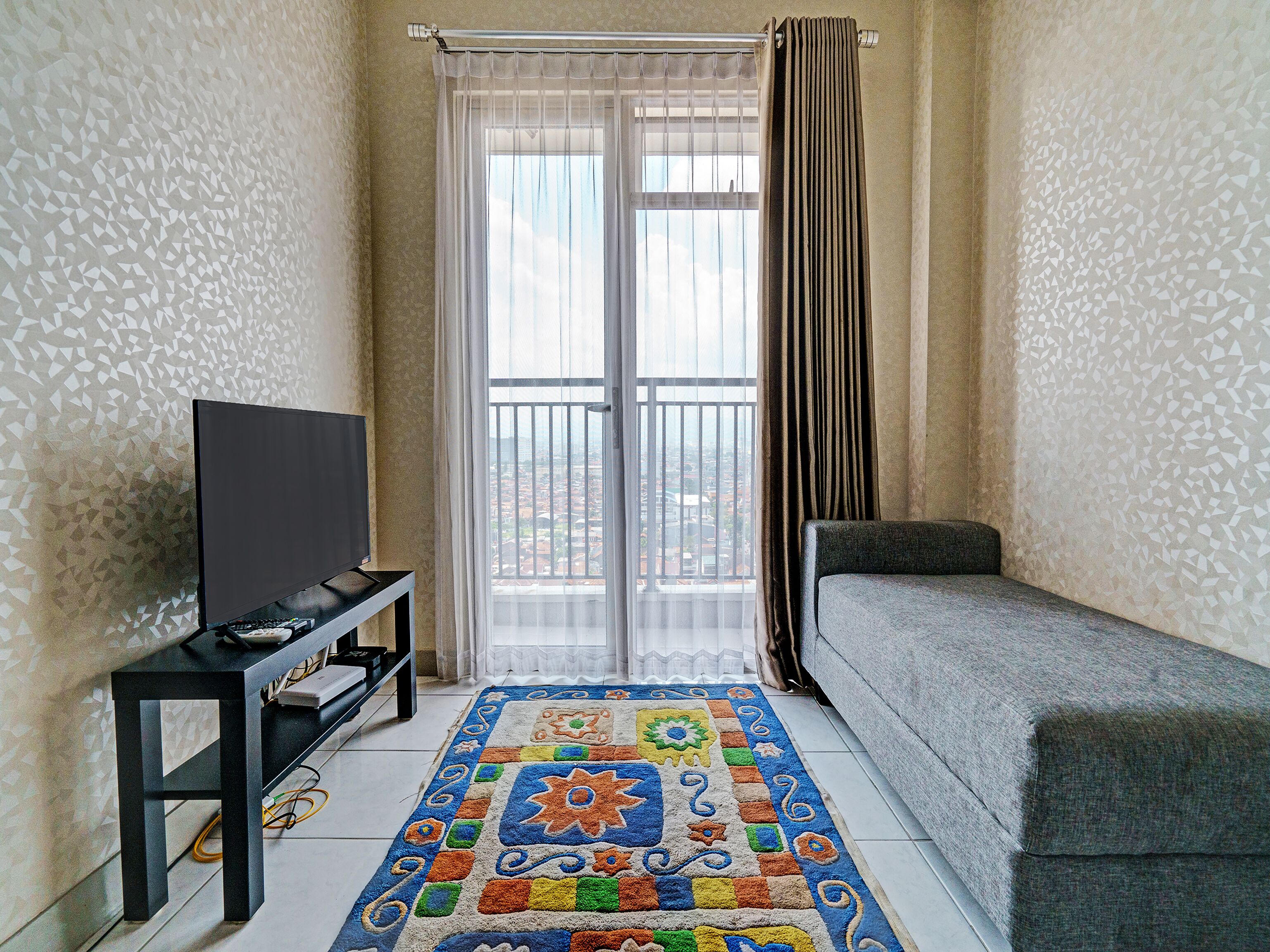 Une petite pièce moderne avec un canapé gris, un téléviseur posé sur un meuble noir, un tapis à motifs et un balcon offrant une vue sur la ville.
