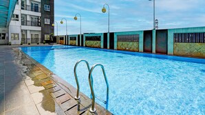 A swimming pool with clear blue water, surrounded by a low wall with a metal railing. There are potted plants along the wall, and a building with balconies is visible in the background. - OYO Life 92548 M-square Apartment By Lins Pro (Bandung)