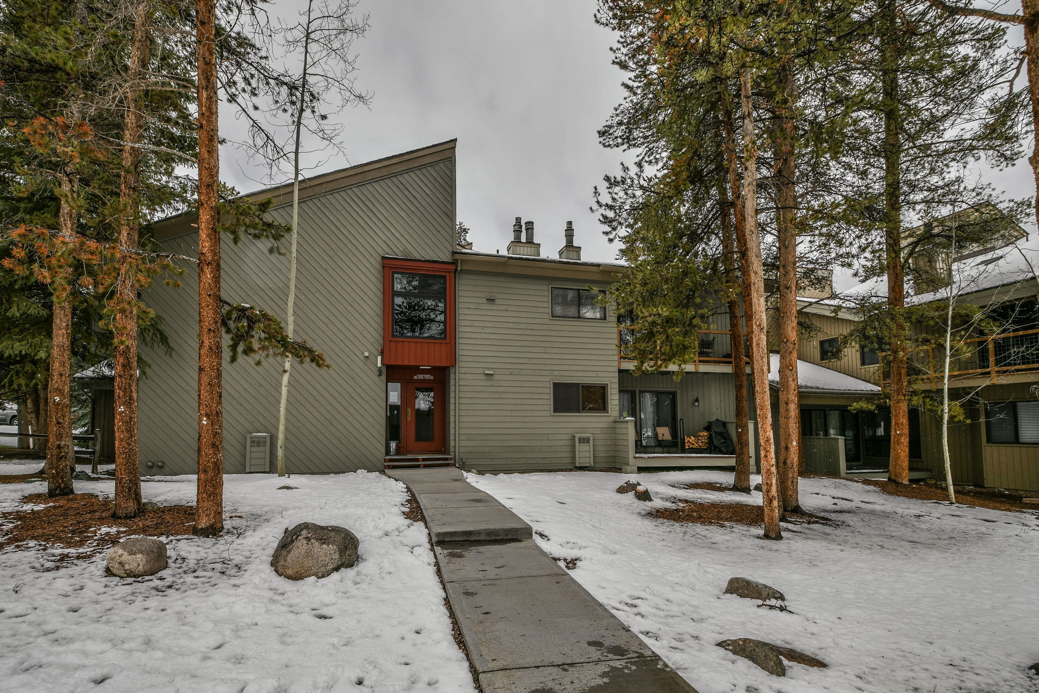 A modern building with a sloped roof and a red door, surrounded by tall trees and a snow-covered ground.