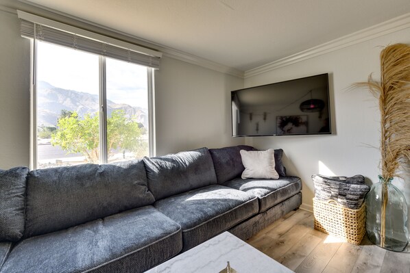 Un salon moderne avec un canapé d’angle gris, un téléviseur à écran plat fixé au mur et une grande fenêtre donnant sur un paysage de montagnes et d’arbres.