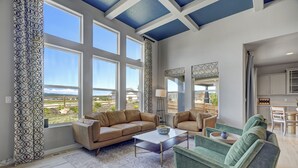 A spacious living room with large windows, a beige sofa, a green armchair, and a coffee table.