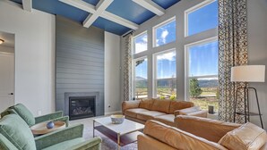 A living room with a fireplace, green and beige sofas, and large windows offering a view of the outdoors.