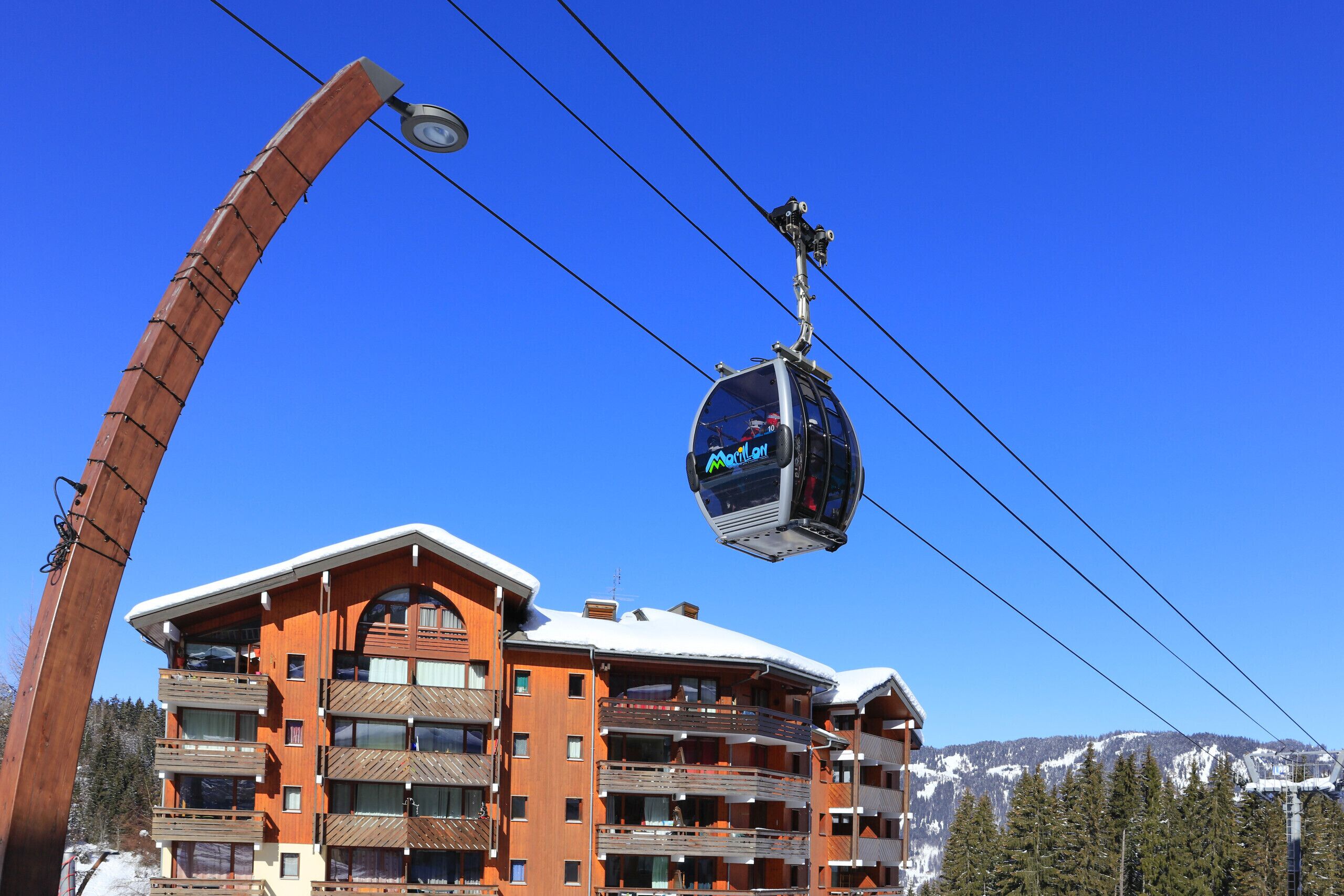 A cable car system with a wooden structure and a building with multiple balconies and a ski slope in the background.