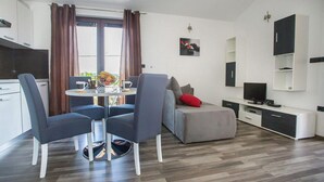 Un salon moderne avec un canapé gris, une table basse et un téléviseur. On y trouve également un coin repas avec une table et des chaises, ainsi qu’une cuisine équipée de placards et dotée d’une fenêtre.