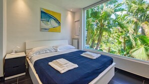 A bedroom with a bed, a nightstand, a window with a view of tropical plants, and a painting on the wall.