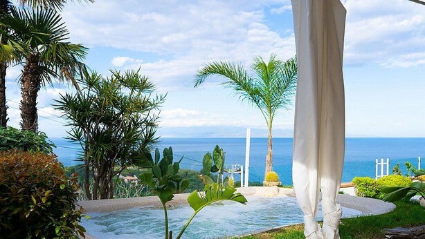 A pool area with a view of the ocean, surrounded by tropical plants and a white structure.