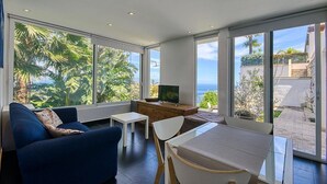 A modern living room with a sofa, a TV on a wooden cabinet, and a dining table. Large windows offer a view of palm trees and a building.