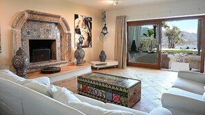 A living room with a tiled fireplace, white sofas, a colorful coffee table, and a view of a mountainous landscape through large glass doors.