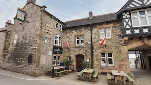 Un edificio de piedra tradicional con un letrero que dice 'Bulls Head Hotel' y 'Marston's'. Hay mesas y bancos al aire libre, y se ve una bandera.