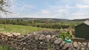 Un muro de piedra con un jardín en primer plano, un cobertizo verde y, al fondo, vistas de colinas y campos ondulantes.