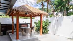 A covered outdoor seating area with a thatched roof, a pool, and a white wall. - Paradise Found (Rawai)