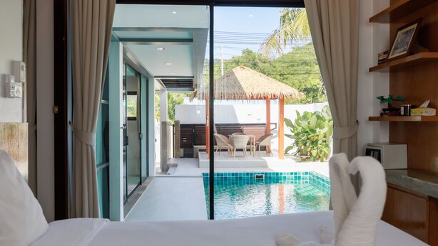 A hotel room with a large bed, a view of a swimming pool, and a dining area with a thatched roof. - Paradise Found (Rawai)