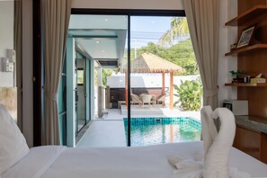 Quarto de hotel com uma cama grande, vista para uma piscina e área de refeições com telhado de palha.