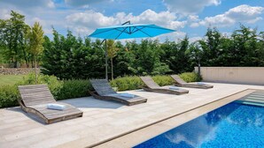 Un espace aménagé au bord de la piscine, avec quatre chaises longues en bois, un parasol bleu et une piscine à l’eau d’un bleu limpide.
