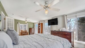 A bedroom with a large bed, a ceiling fan, a dresser, a TV, and a window with a view of trees. - Always Somerset (Colorado Springs)