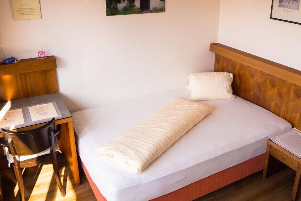 Ein ordentlich eingerichtetes Schlafzimmer mit einem Bett, Kissen, einem Holz-Kopfteil, einem Schreibtisch mit Papieren und einem Stuhl.