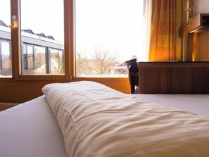 Une chambre avec une grande fenêtre, un lit avec une literie blanche et une table de chevet en bois.