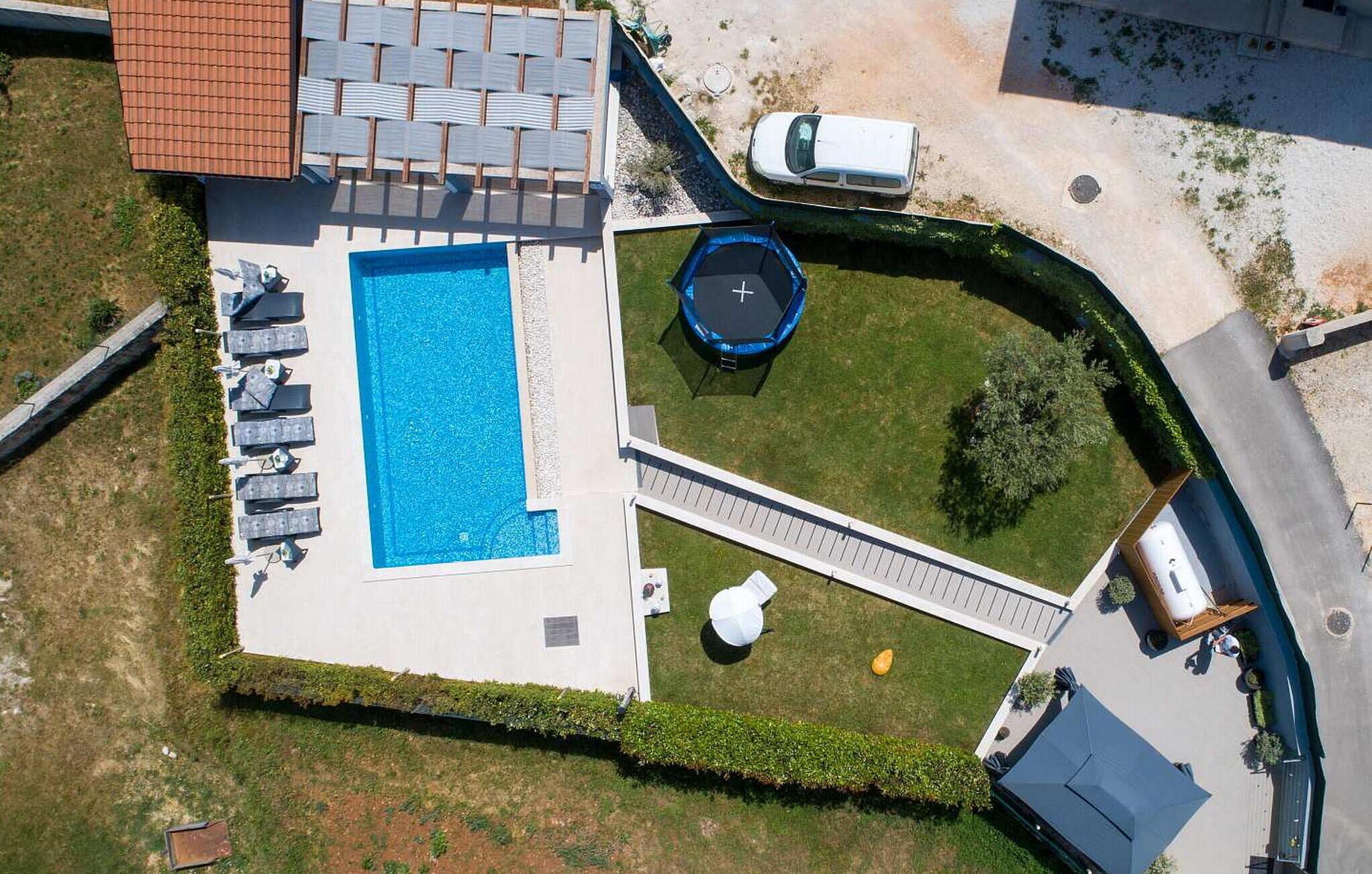 An aerial view of a property with a swimming pool, sun loungers, a trampoline, and a covered outdoor seating area.
