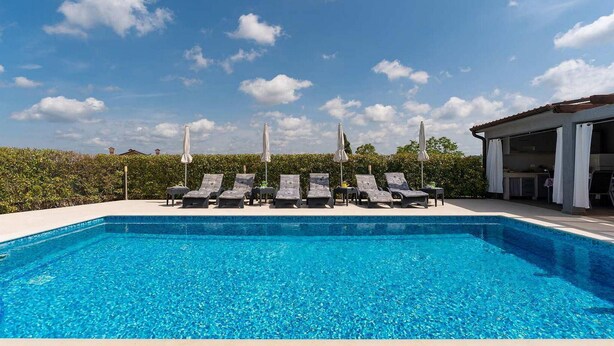 En klarblå pool med solstolar och parasoller, omgiven av grönska och en byggnad.