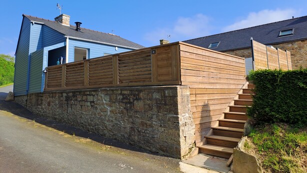 A two-story house with a stone wall and wooden fence, a clear blue sky, and a road in the foreground.