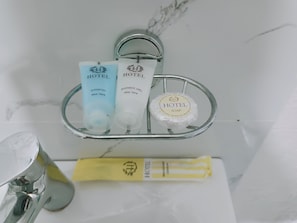 A bathroom with a glass shelf holding hotel toiletries including shampoo, shower gel, and soap. - Nomad Inn Guest House (Bishkek)