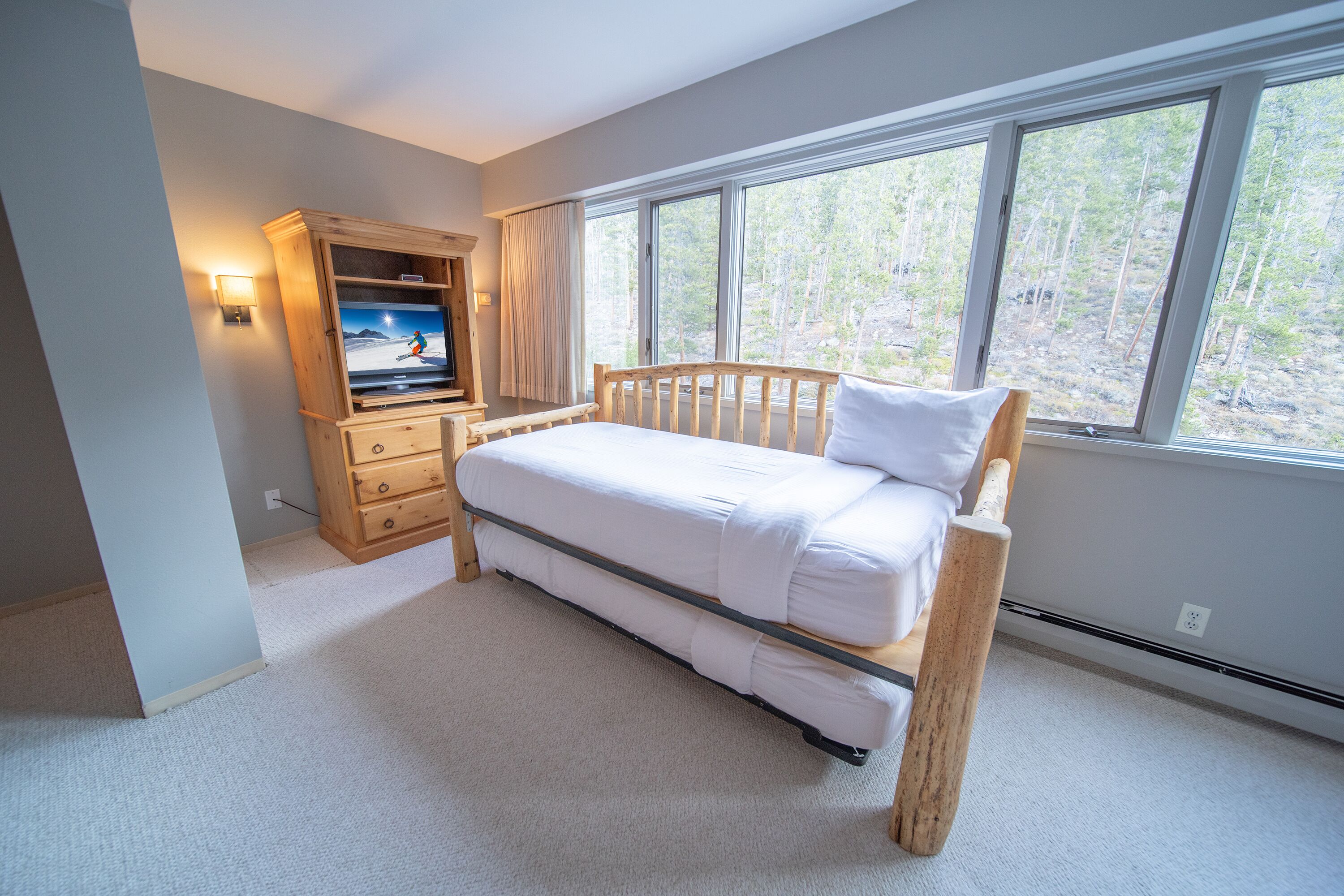 Quarto com cama de madeira, televisão sobre um móvel de madeira e janela com vista para a floresta.