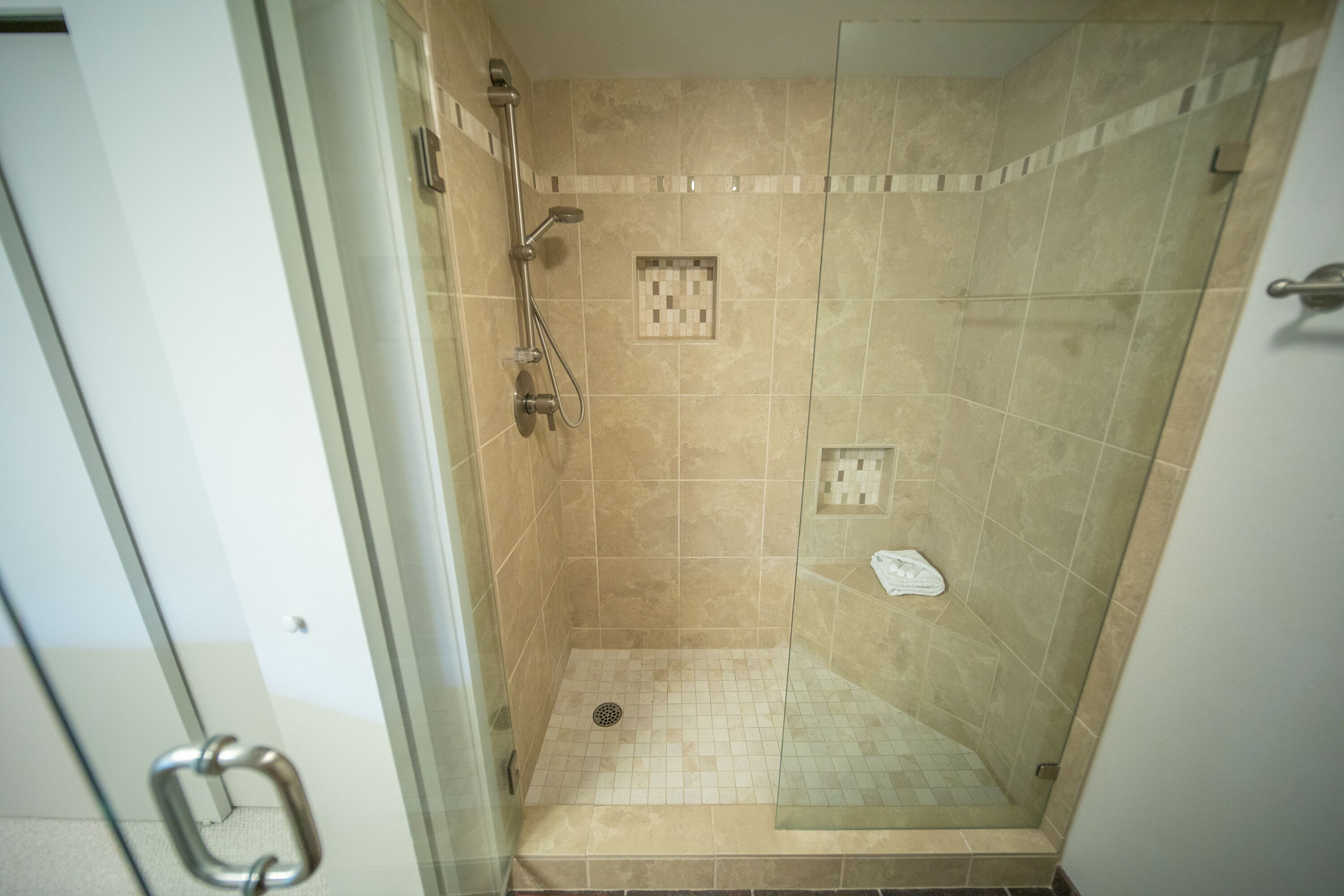 Een douchecabine met glas, bekleed met beige tegels, voorzien van een handgreep en een klein handdoekenrek.