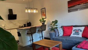 A modern living room with a grey sofa, a wooden coffee table, and a red rug. There is a kitchen area with a bar and a TV mounted on the wall. - Studio Apartment mit Balkon, am See mit Privat Badestrand. Haustierfreundlich! (Afritz)