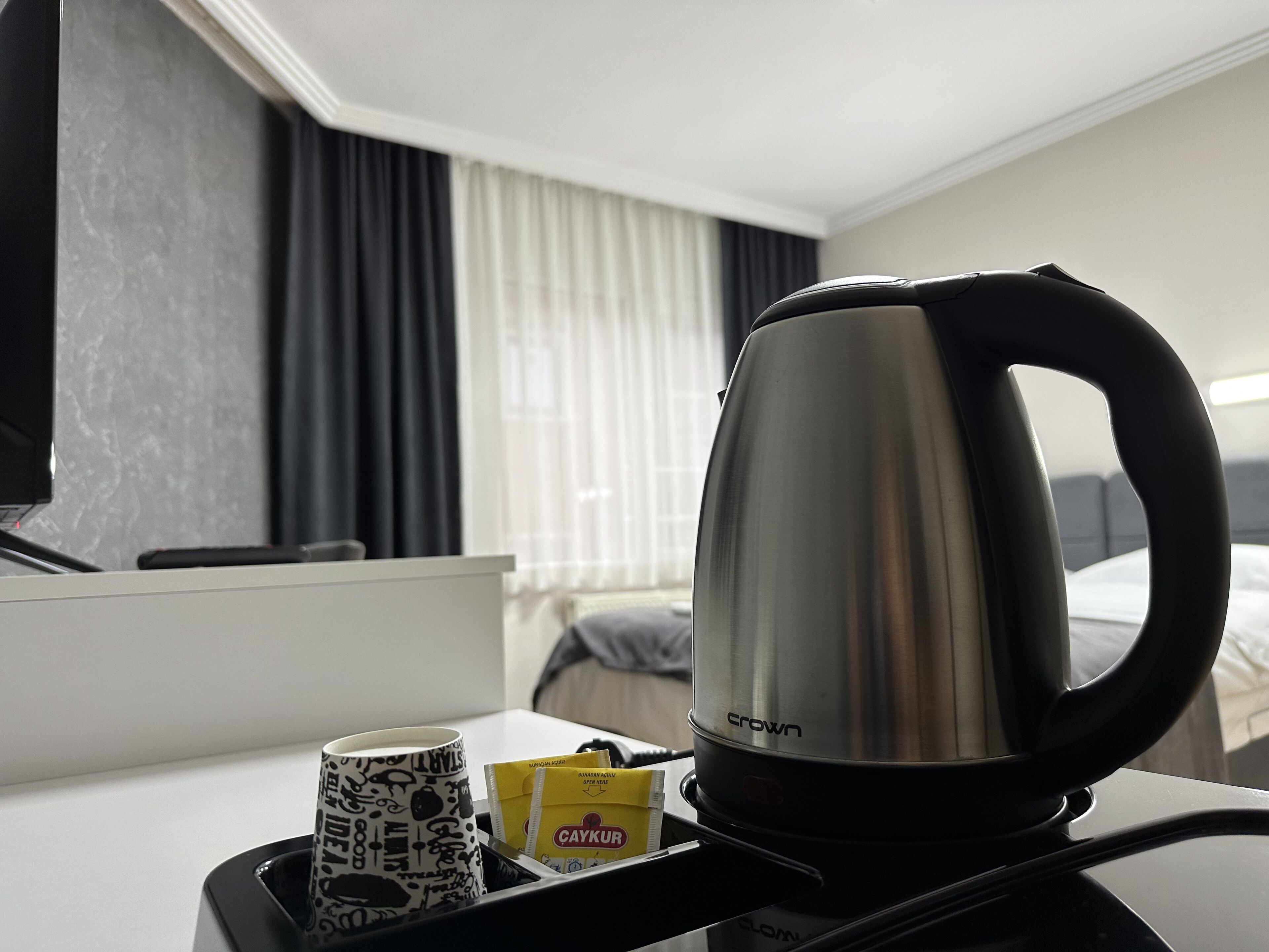 A hotel room with a bed, a window with curtains, a coffee maker, a mug, and a tray with a bag of snacks.