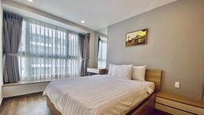 A hotel room with a large bed, a bedside table, a nightstand, a window with curtains, and a painting on the wall.