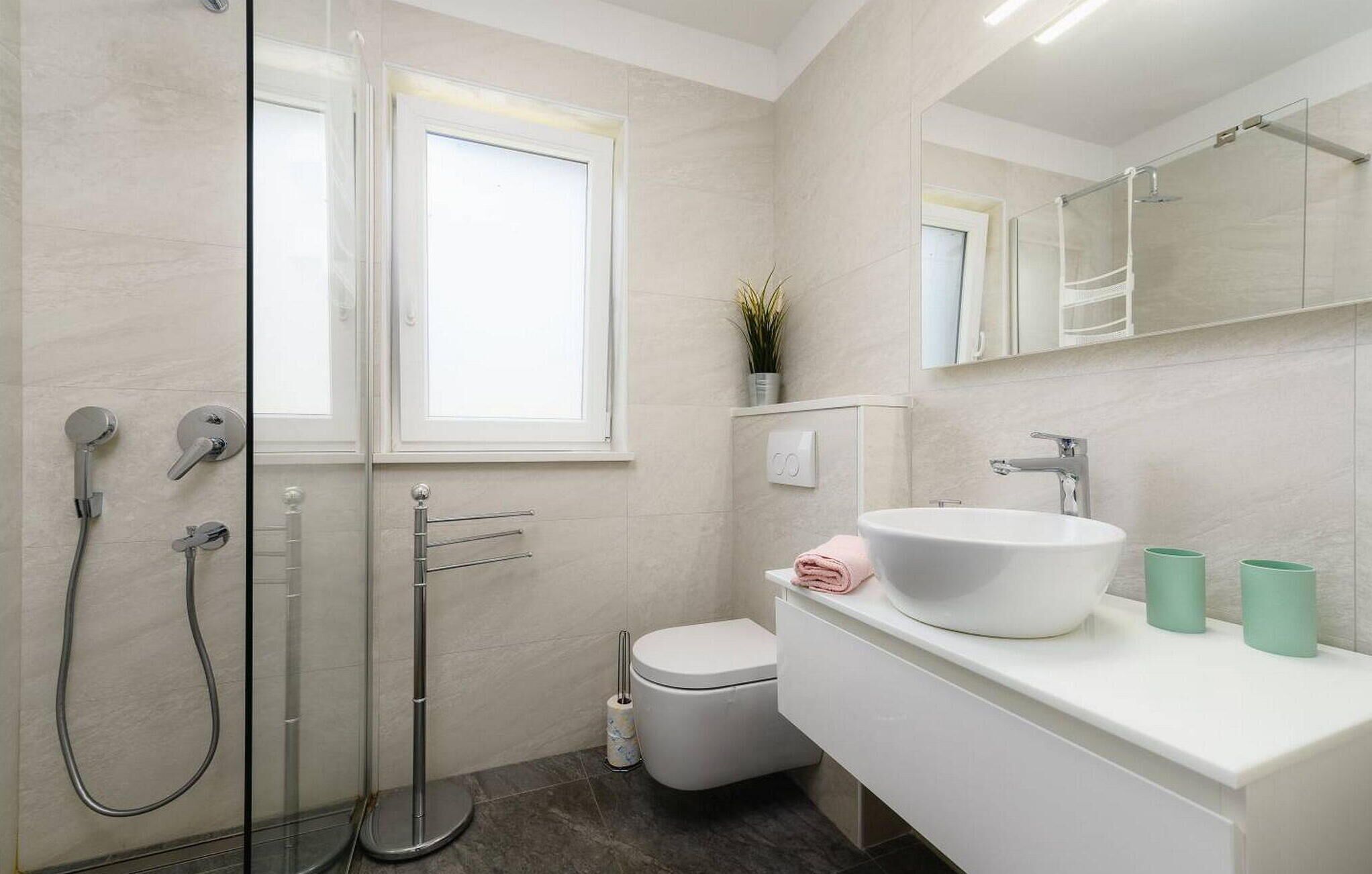 A modern bathroom with a glass shower enclosure, a white toilet, a large sink with a mirror above, and a window providing natural light.