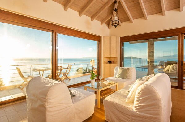Ein geräumiges Zimmer mit großen Fenstern, die einen Pool und das Meer zeigen, mit heller Möbel und Holzboden.