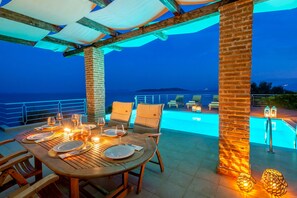 An outdoor dining area with a pool in the background, wooden tables, and chairs. - Seafront 'Karavi Villa': 270° Views & Private Sea Access (Sivota)