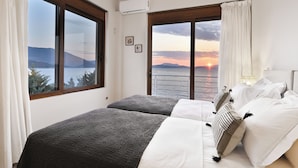 A bedroom with two beds, large windows with curtains, a view of the sunset, and a ceiling light. - Beachfront 'Karavi Villa': 270° Views & Sea Access (Sivota)