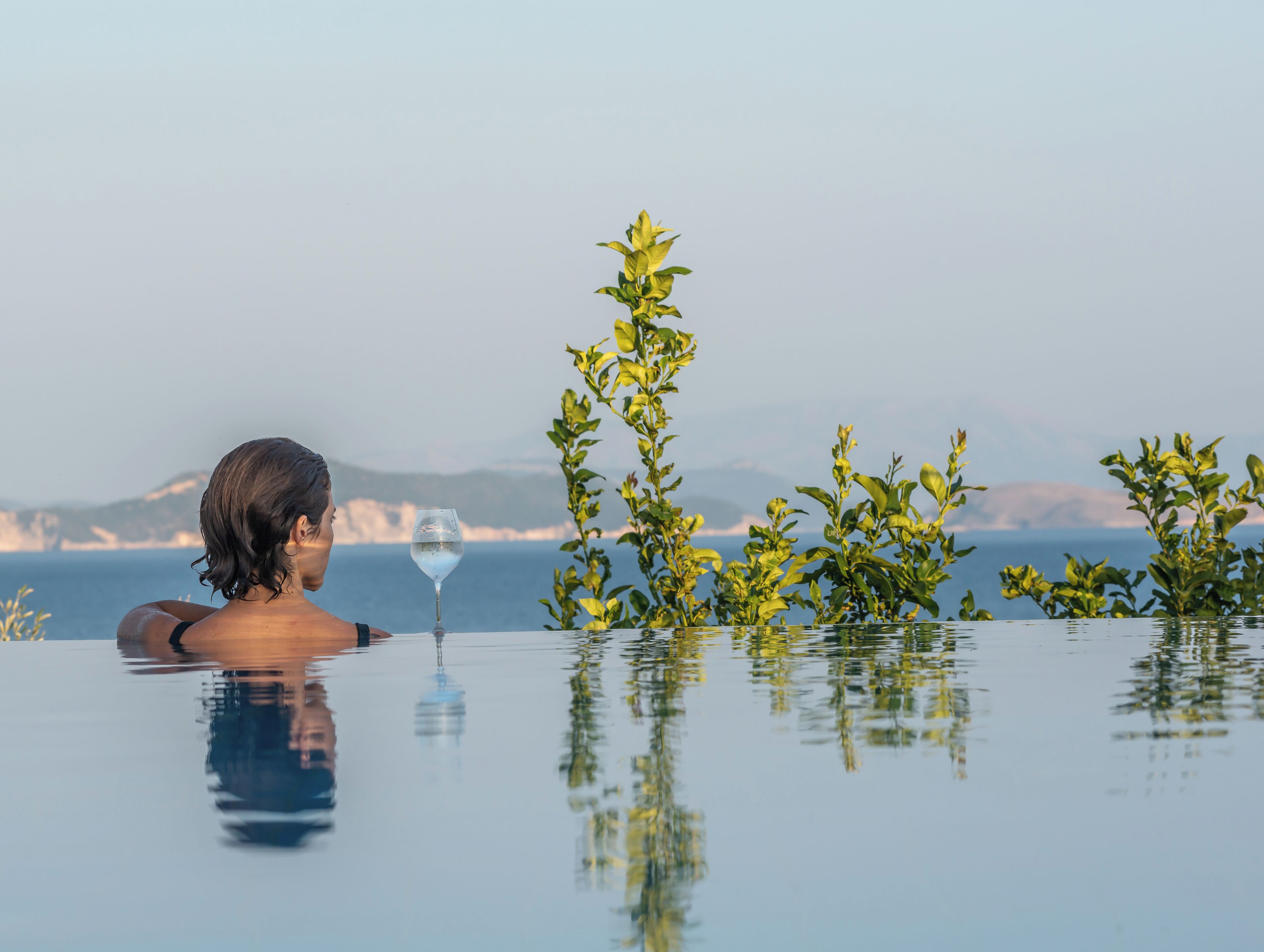 Une personne se détend au bord d’une piscine, un verre d’eau à la main, entourée de verdure et avec une vue sur la mer.