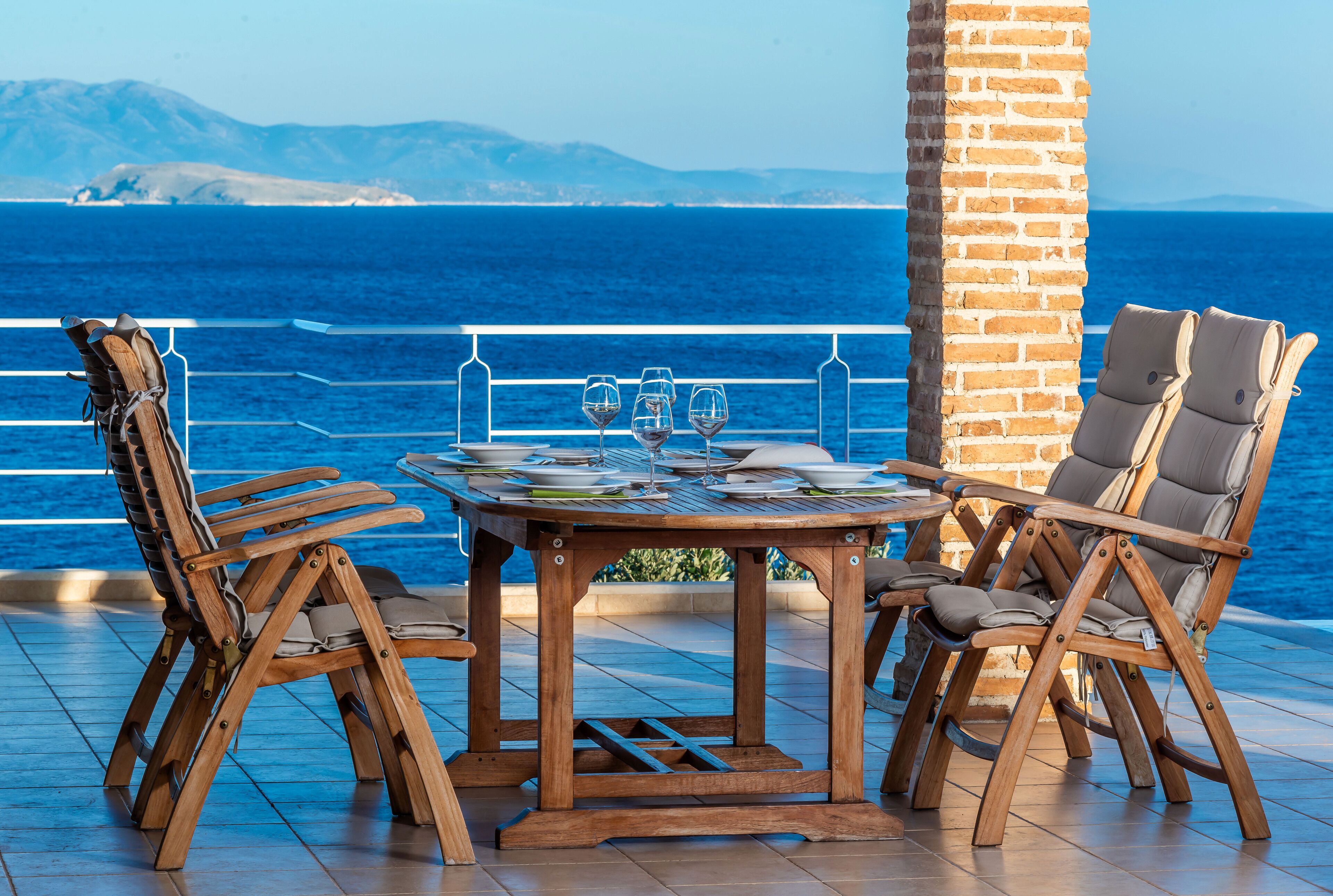 A wooden table with four chairs, set on a terrace with a view of the sea and mountains.