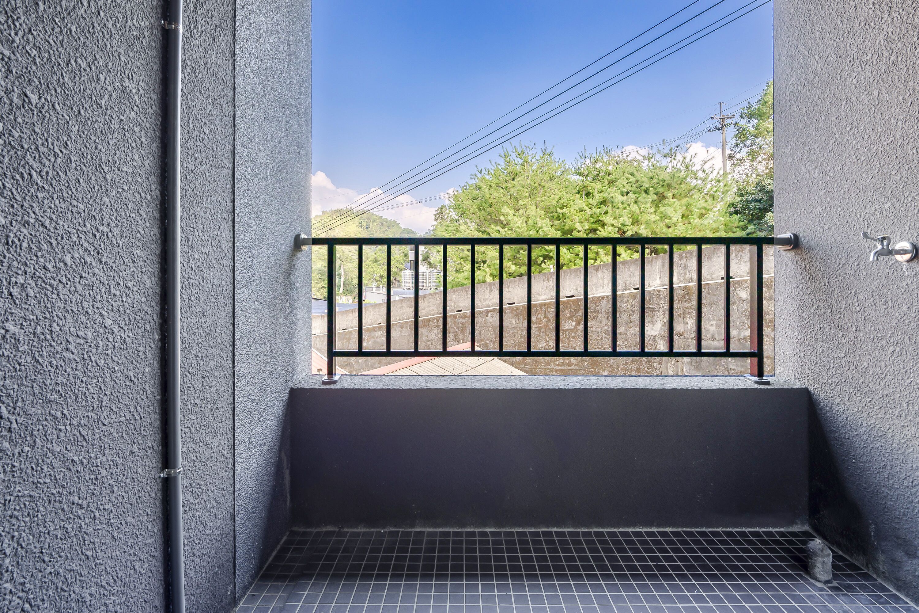 Un balcón con barandilla de metal, piso de baldosas y vistas a vías de tren y follaje verde.