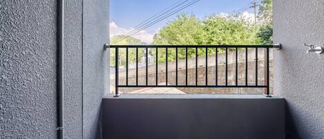 Un balcón con barandilla de metal, piso de baldosas y vistas a vías de tren y follaje verde.