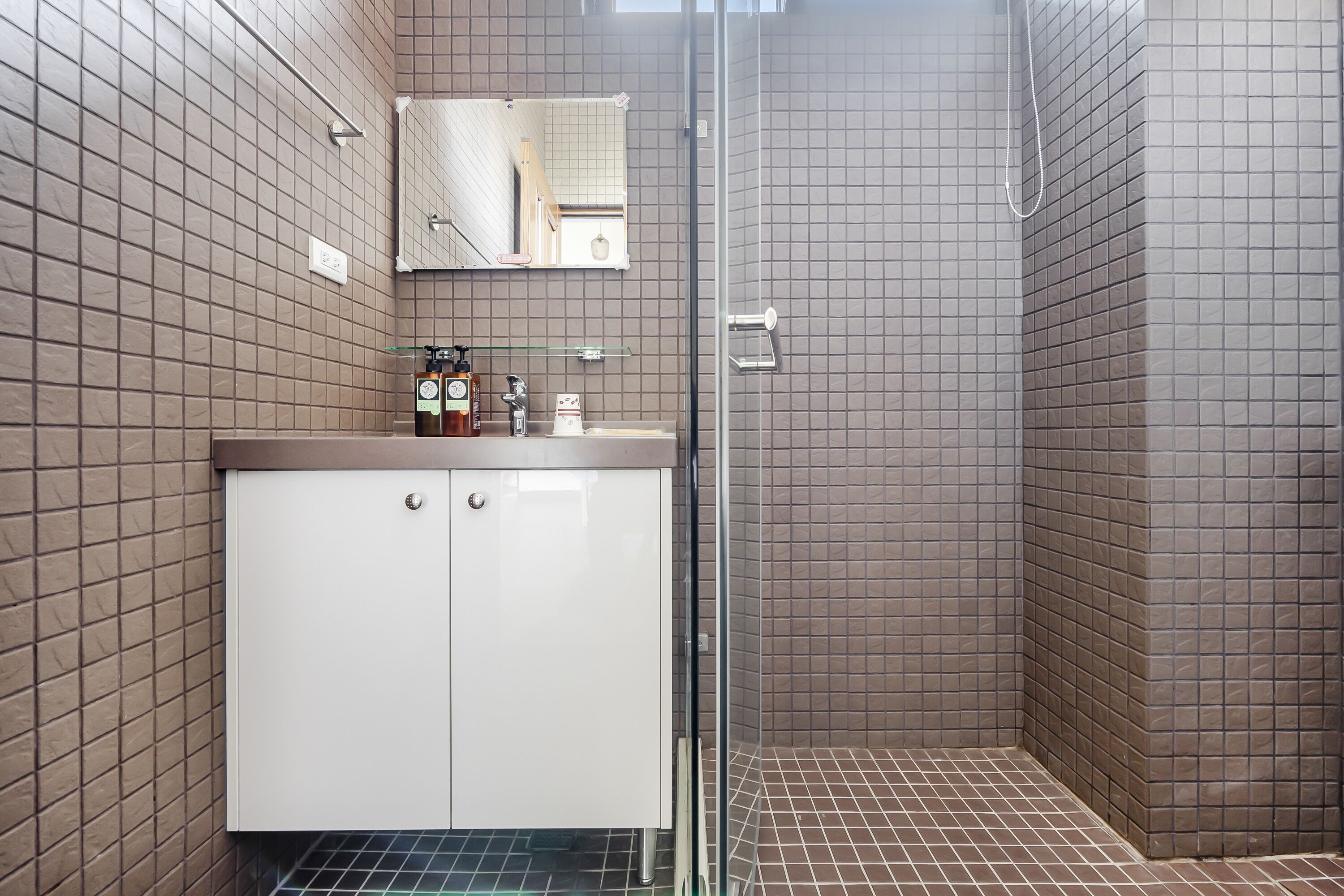 A modern bathroom with a glass shower enclosure, a white vanity with a mirror, and brown tiled walls.